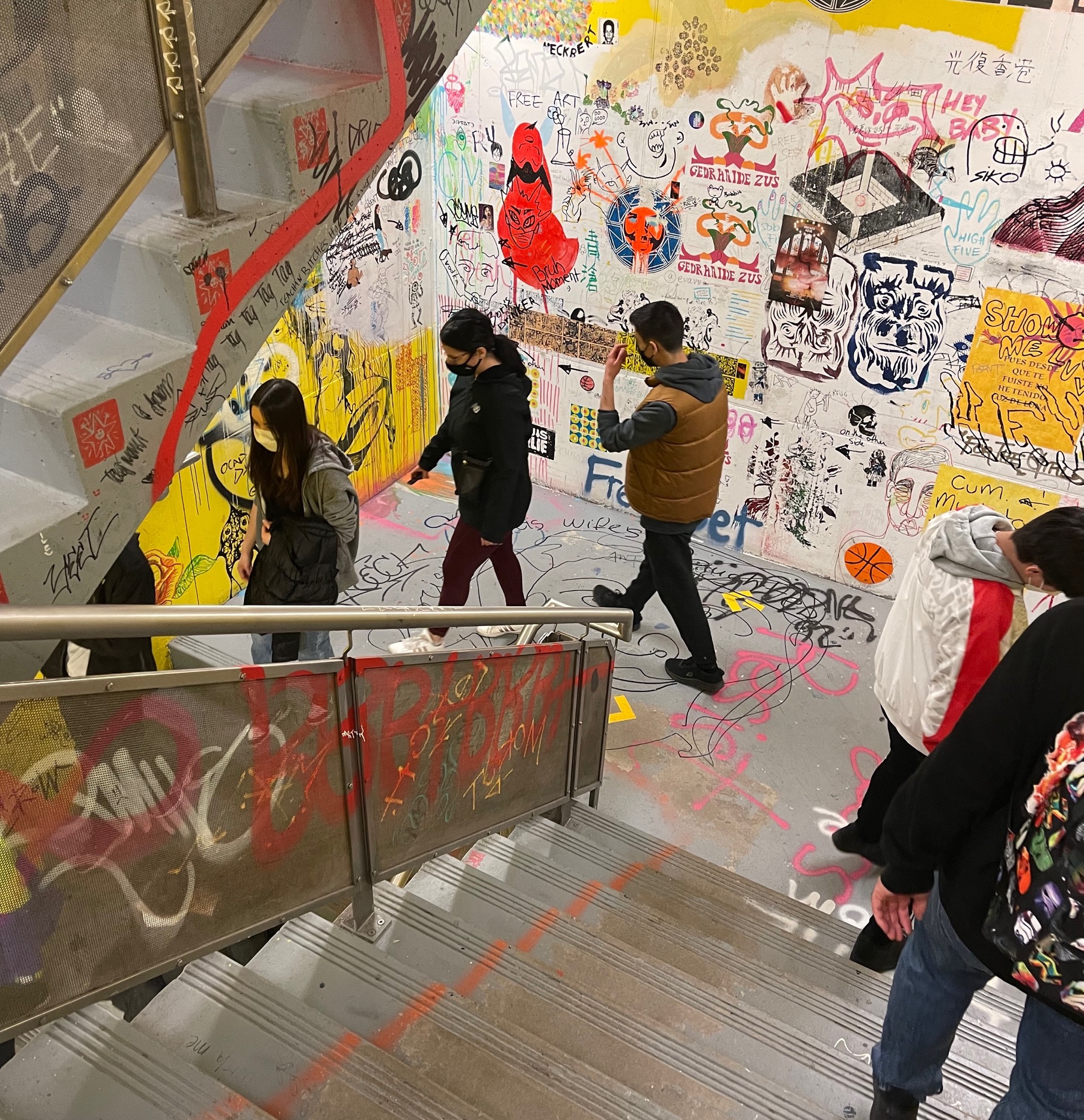 Masked tour group walking through a colourful, graffiti-covered staircase.