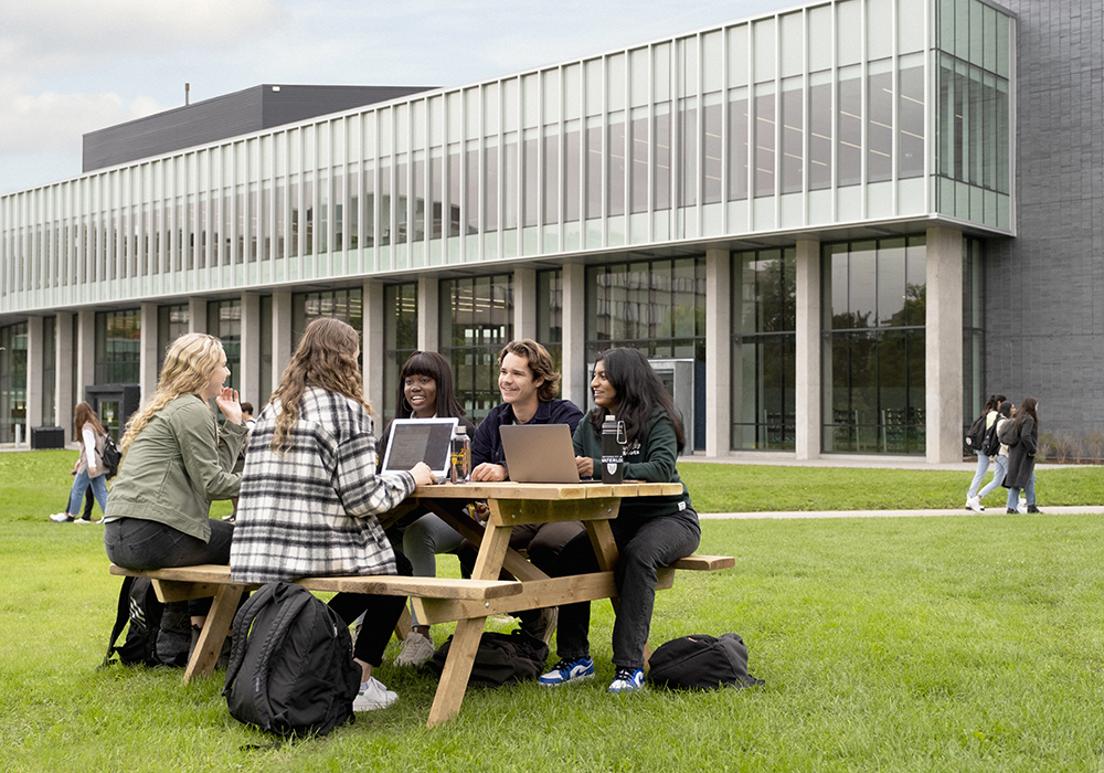 Students sitting outside at a picnic table in front of the Student Life Centre building on campus