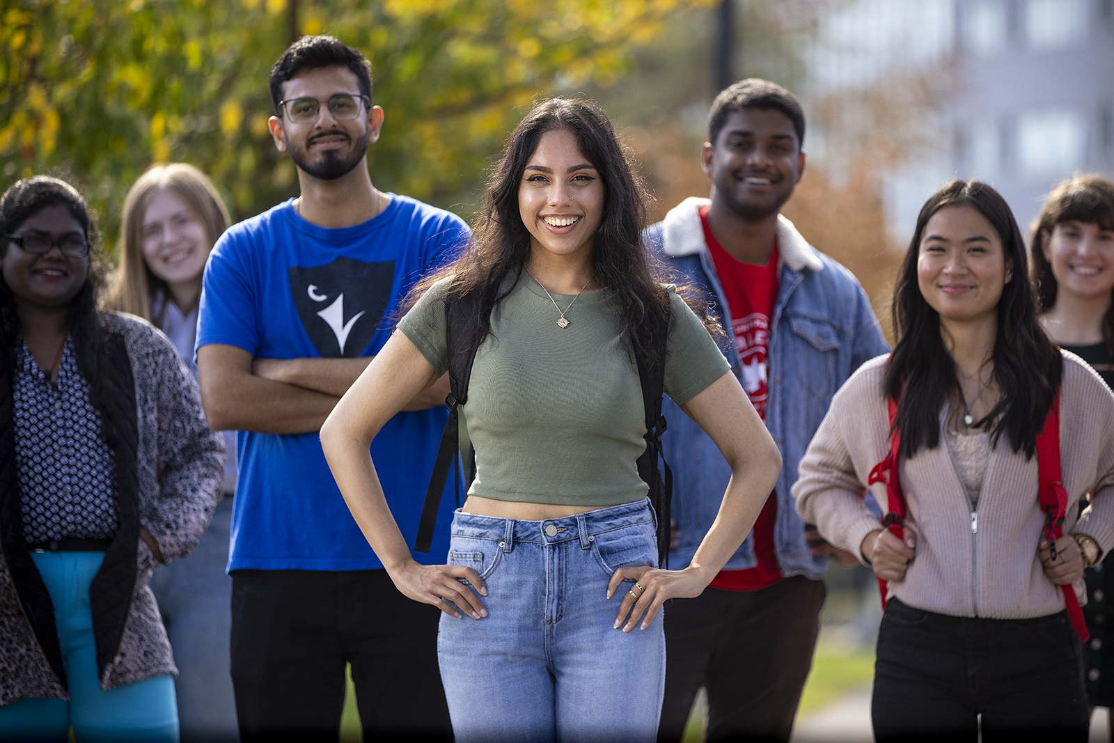 Group of smiling students outside on campus, trees in background, all standing facing the camera, young woman centre front has hands on hips
