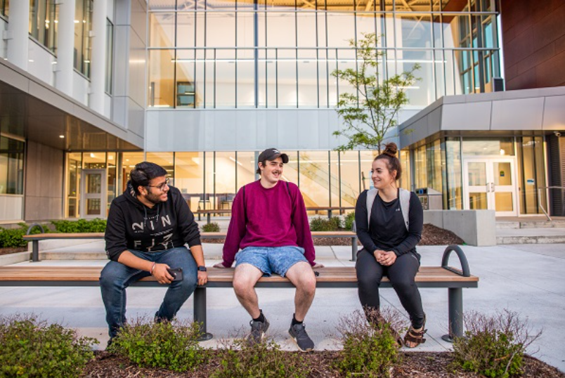 Photo of 3 students sitting on a bench outside the Nova building at Lambton's main Sarnia campus