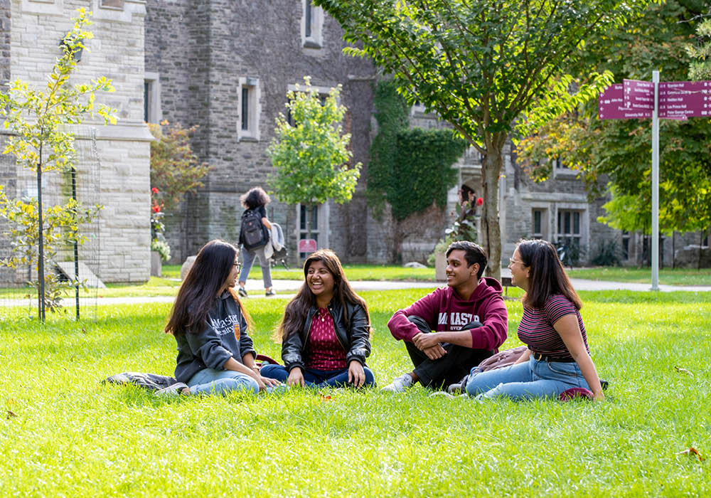 Four young adults sit together on a patch of grass on a summer day at McMaster University in Hamilton, Ontario, Canada.