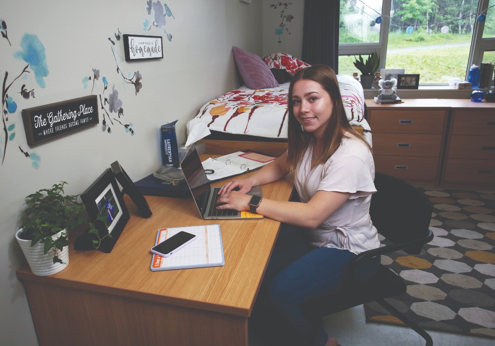 A Laurentian student sitting at her desk in Residence, with her bed in the background.