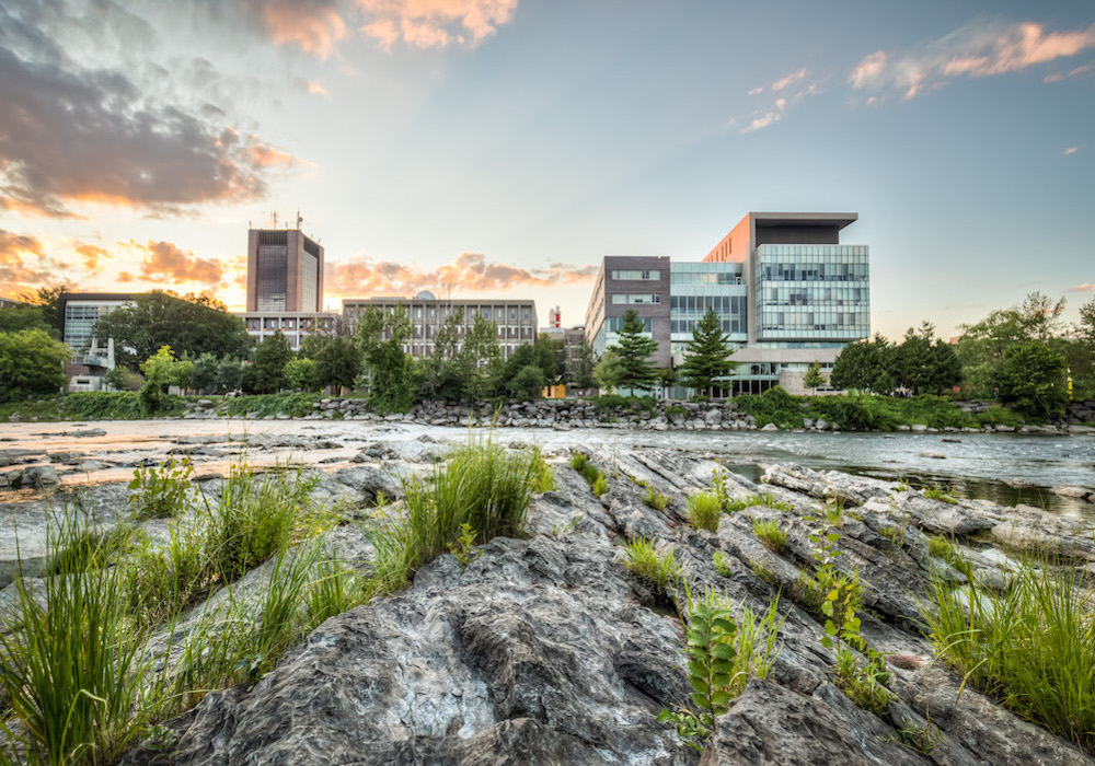 View of buildings, Carleton campus from other side of Rideau river - river, rocks, green grass and trees, clouds near sunset, blue sky