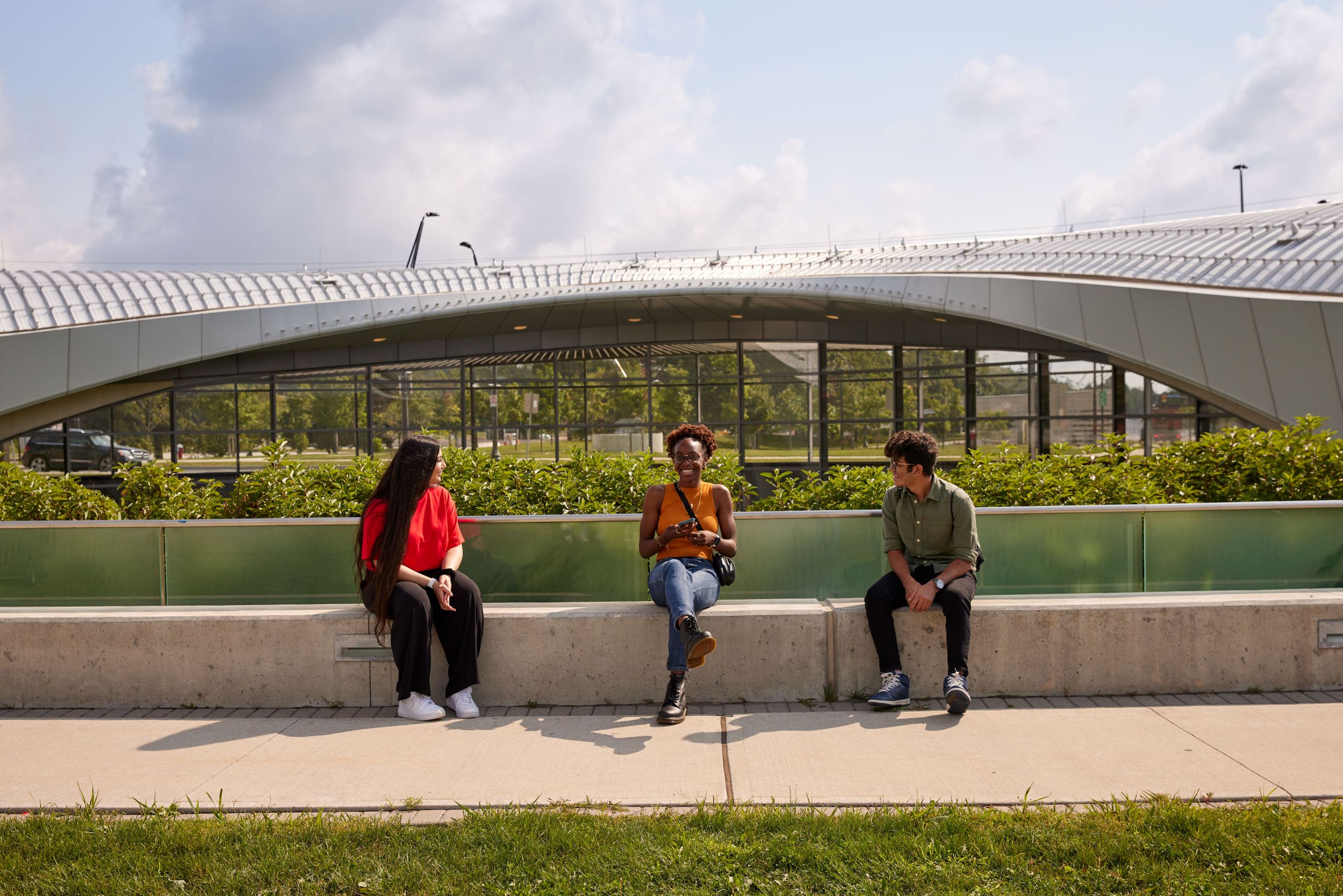 Three students sit smiling and talking in front of a glass-paneled subway station.